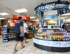 two people walking in front of a store with bottled drinks on the counter and shelves