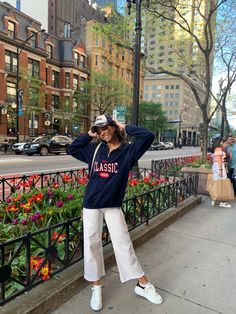 a woman is standing on the sidewalk in front of flowers and buildings with her hands behind her head