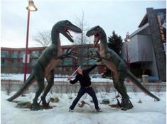 a man standing next to two fake dinosaurs in front of a building with snow on the ground