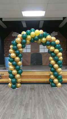 an arch made out of gold and green balloons in front of a wooden table with chairs