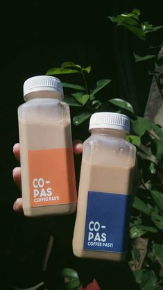 two bottles of coffee sit next to each other in front of some plants and leaves