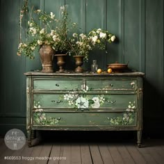 an old dresser with flowers and vases on it in front of a green wall