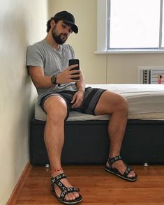 a man sitting on a bed looking at his cell phone while wearing sandals and a hat