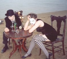 two people dressed up in costumes sitting at a table on the beach with wine bottles