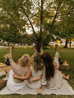 three women sitting on a blanket under a tree and holding wine glasses in the air