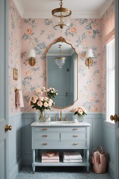 a pink and blue bathroom with floral wallpaper on the walls, a gold framed mirror, an antique style sink and two vases filled with flowers