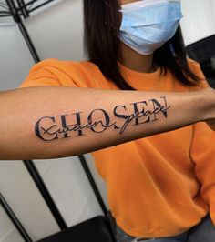 a woman wearing a face mask and holding up her arm with the word chosen tattooed on it