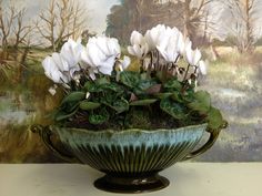 a green bowl with white flowers in it on a table next to a wall painting