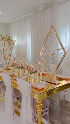 a dining room table set up with gold and white chairs, pink flowers and candles