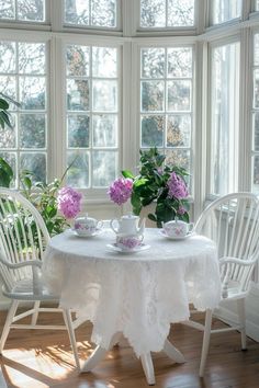 two white chairs and a table with purple flowers on it in front of large windows