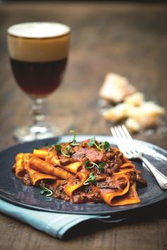 a black plate topped with pasta and meat next to a glass of wine on a table