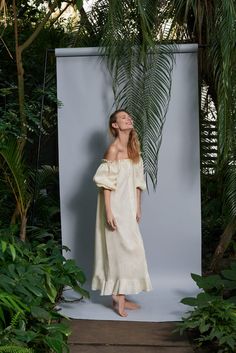 a woman standing in front of a white backdrop with palm trees and plants around her