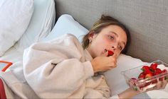 a woman laying in bed eating strawberries with a bowl of fruit on her lap