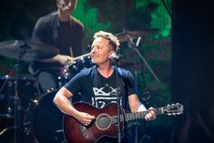 a man holding a guitar while standing in front of a microphone and another person behind him