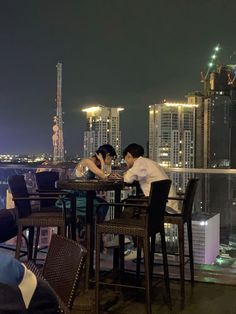 two people sitting at a table on top of a building with city lights in the background