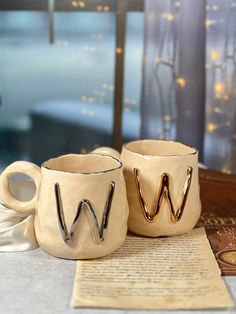 two coffee mugs sitting next to each other on top of a table near a window