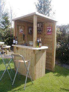 a wooden bar with chairs around it on the grass in front of some bushes and trees