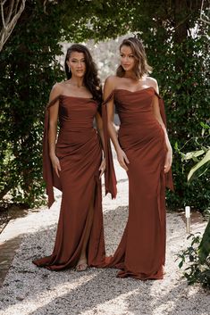 two women in long brown dresses standing next to each other on a path with trees and bushes behind them