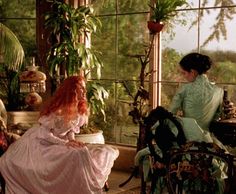 two women dressed in period clothing sitting at a table with plants and potted plants