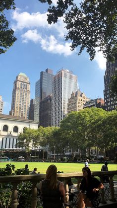 two people sitting on a park bench in front of tall buildings and green grass with trees