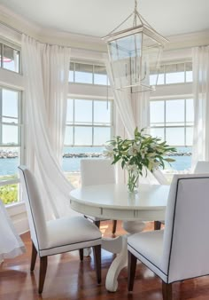 a dining room table with white chairs and a vase on top of it in front of two large windows