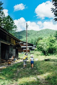 two people walking down a dirt road in front of wooden buildings with mountains in the background