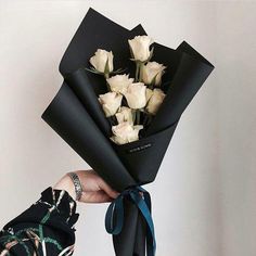 a person holding a bunch of white roses wrapped in black paper and tied with a blue ribbon