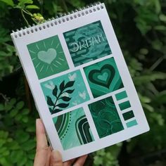 a hand holding up a spiral notebook with images of hearts and leaves on it in front of green foliage