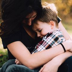 a woman holding a small child in her arms while sitting on the ground with grass behind them