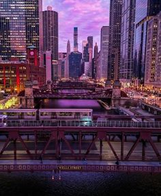 a train traveling over a bridge in the middle of a city at night with lights on