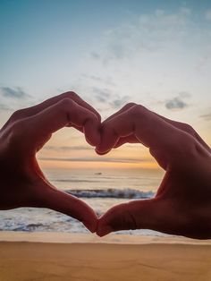 two hands making a heart shape with the sun setting in the background at the beach