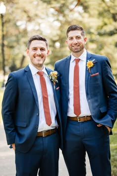 two men in suits standing next to each other on a sidewalk with trees in the background