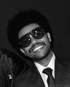 a man wearing sunglasses and a suit smiling at the camera with another man in the background