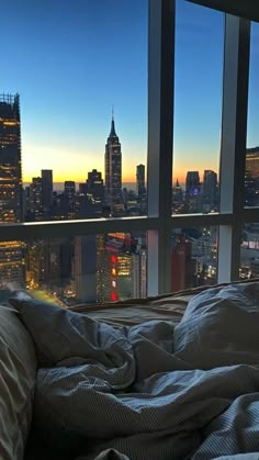 an unmade bed in front of a large window overlooking the city at night time