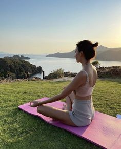 a woman sitting on a yoga mat in the middle of a grassy field with water and mountains in the background