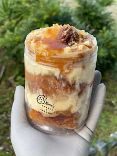 a hand holding a jar filled with desserts on top of green grass and trees