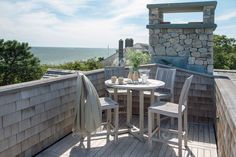 an outdoor table and chairs on a wooden deck overlooking the ocean with a stone chimney in the background