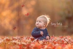 a baby sitting on the ground surrounded by leaves and looking up at something in the air