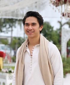 a young man wearing a white shirt and tan shawl smiles at the camera while standing in front of a chandelier