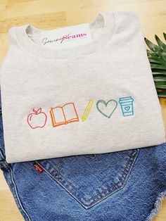 Brighten a teacher’s day with our Teacher Elements Embroidered T-Shirt! This delightful t-shirt features an array of teacher icons embroidered in the vibrant colors of the rainbow, creating a simple yet effective design that celebrates the passion and dedication of educators. Crafted from high-quality, soft, and breathable fabric, this t-shirt offers all-day comfort and durability. It’s perfect for wearing to school, casual outings, or lounging at home, making it a versatile addition to any teac Teacher Shirt Designs Ideas, Cute Teacher Sweatshirts, Teacher Hand Embroidery Designs, Teacher Hand Embroidery, Embroidery For Teachers Gift Ideas, Embroidered Teacher Gifts, Embroidery Teacher Gifts, Teacher Inspired Outfits, Future Teacher Wallpaper