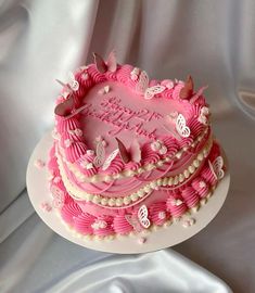 a pink heart shaped birthday cake on a white plate with pearls and butterflies around it