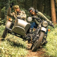 two men riding on the back of a motorcycle in the woods with trees behind them