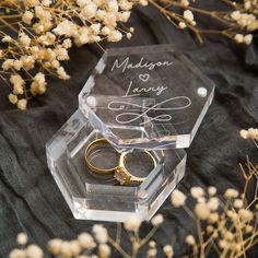 two wedding rings are placed in an acrylic box on a black cloth with dried flowers