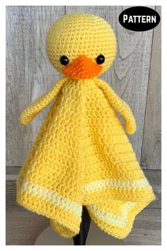 a crocheted yellow duck with black eyes is sitting on a wooden stand and has a knitted blanket around it