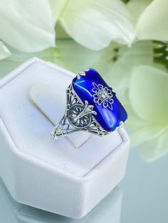 Sapphire Blue Glass Sterling Silver Ring Grace Design#D233 This is a lovely antique Edwardian era inspired filigree ring. This gorgeous ring is created in stunning sapphire blue glass. The glass is embellished with a delicate floral filigree. The top of the ring holds a center set 1mm round cut gemstone (Choose from white Cubic Zirconia, lab created white Moissanite, or Natural/Genuine White Diamond). The blue glass is 18mm x 19mm. The ring sits 18mm north-south and 10mm east-west on the finger. Sapphire Filigree Ring For Wedding, Sapphire Filigree Rings For Gift, Sapphire Rings With Filigree Detail For Gift, Sapphire Filigree Rings As A Gift, Sapphire Rings With Filigree Detailing, Perfect As A Gift, Heirloom Blue Filigree Ring For Weddings, Ornate Intaglio Ring For Wedding, Ornate Intaglio Wedding Rings, Ornate Blue Filigree Ring In Sterling Silver