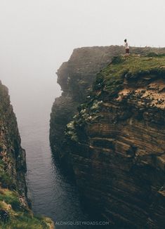 a person standing on top of a cliff next to the ocean with fog in the air