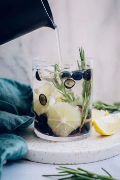 a person pouring olives into a glass with lemon wedges and rosemary sprigs