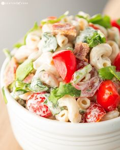 a white bowl filled with pasta salad on top of a wooden table next to tomatoes and lettuce