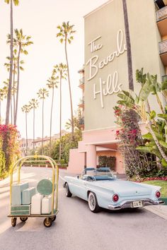 an old blue car parked in front of a hotel with palm trees on the side