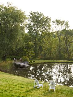 two lawn chairs sitting on top of a lush green field next to a lake and dock